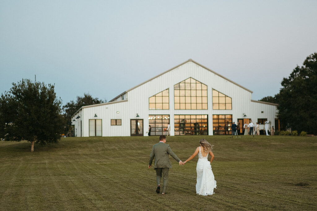 bride and groom walking hand in hand at lakeside wedding venue while guests have cocktails during cocktail hour