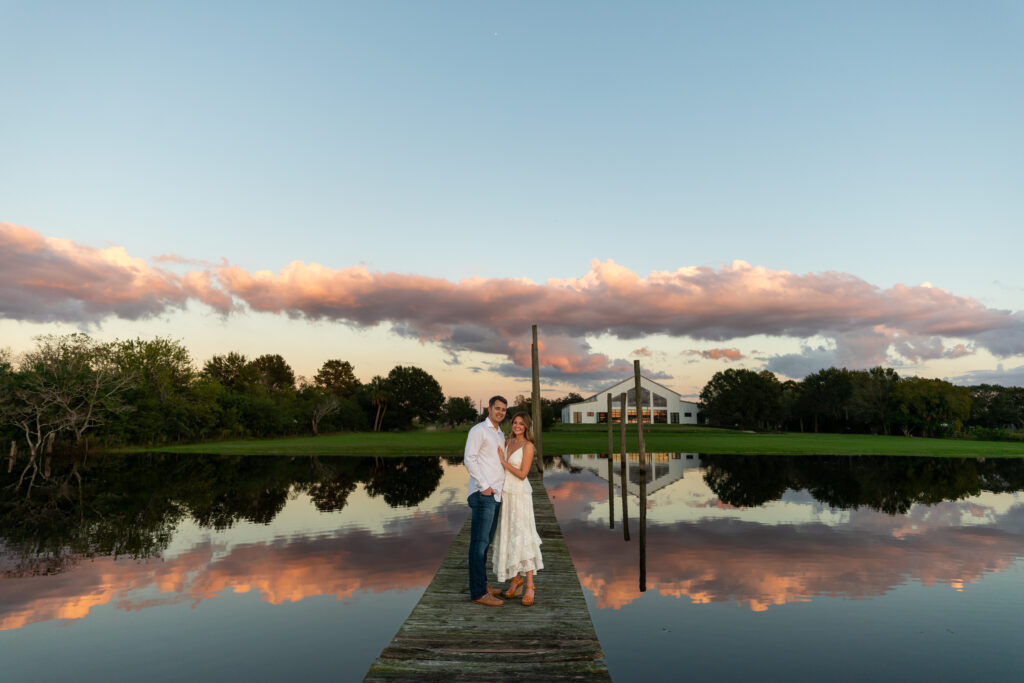 Wedding venue in the distance with a man and woman standing on the dock surrounded by pink clouds reflecting in the lake