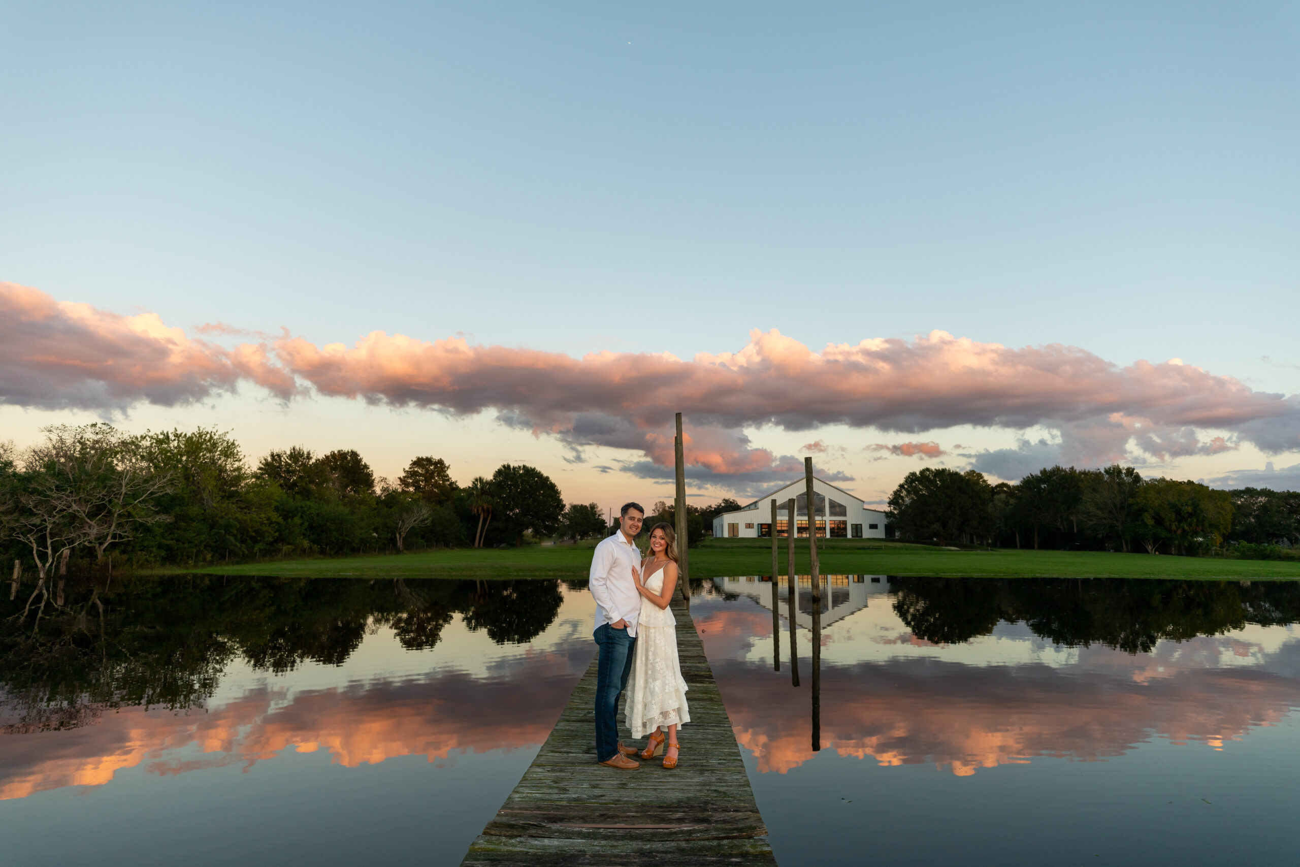 Man and woman standing on a dock on Lake Toho with wedding venue in the background and clouds and sunset reflecting in the water.
